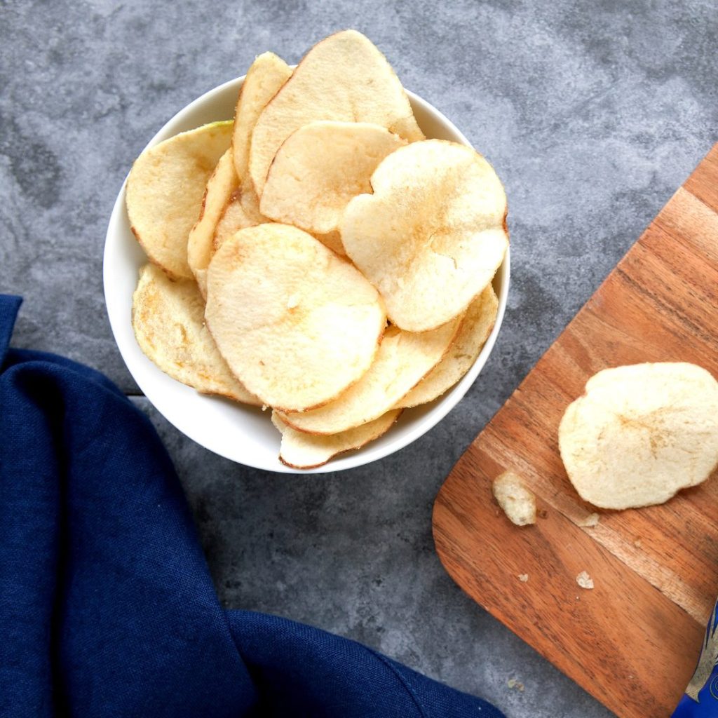 A bowl full of Mackies crisps, sitting next to a wooden chopping board.